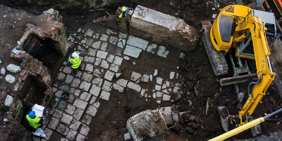 Två arkeologer vid en grävmaskin på en utgrävningsplats där man hittat en gammal husgrund.